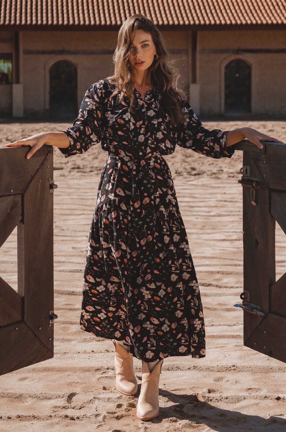 Woman in a floral dress standing in front of wooden gates with a rustic building in the background.