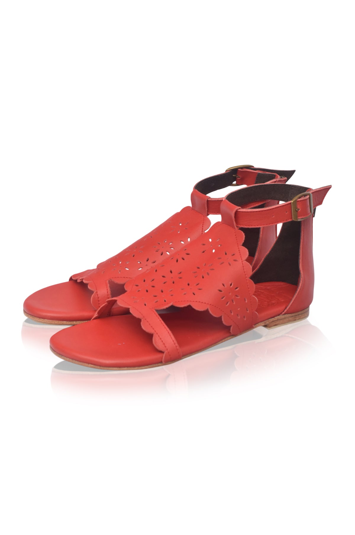 A pair of red flat sandals with scalloped cutout detailing on the toe straps and adjustable ankle straps with small gold-tone buckles, shown on a white background with a soft reflection.