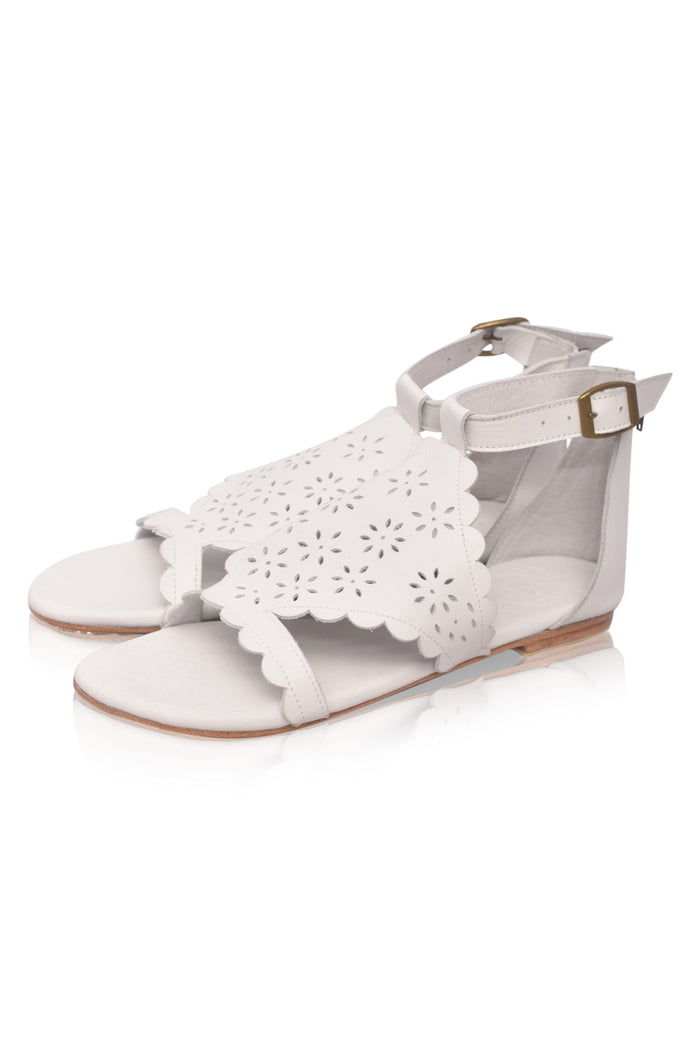 White flat sandals with scalloped laser-cut straps, an adjustable ankle strap, and a small gold buckle, shown on a white background.