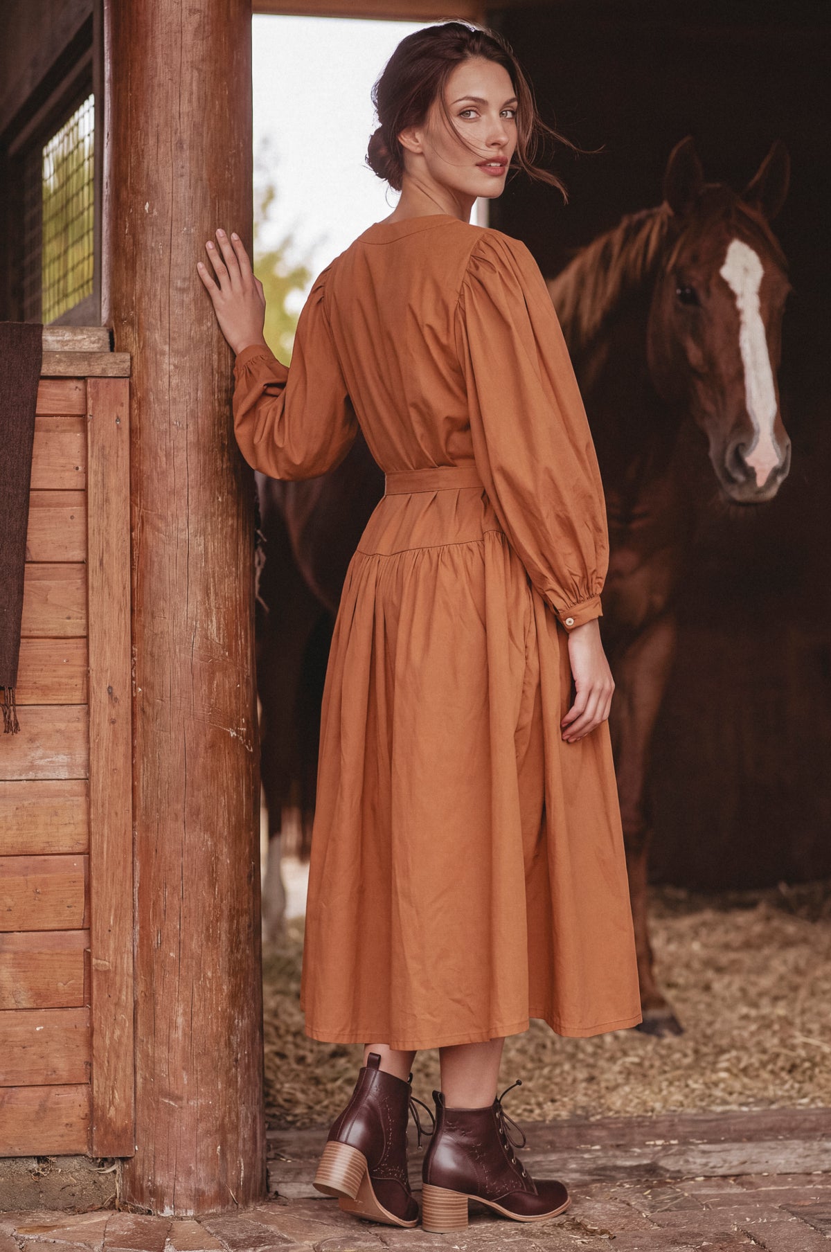A woman wearing a rust-orange long-sleeve midi dress with puff sleeves and a matching belt, standing in a rustic barn next to a wooden post with a brown horse in the background.