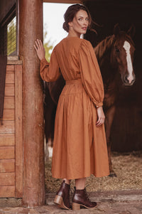 A woman wearing a rust-orange long-sleeve midi dress with puff sleeves and a matching belt, standing in a rustic barn next to a wooden post with a brown horse in the background.