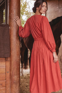 A woman wearing a rust-orange long-sleeve midi dress stands in a rustic barn, leaning against a wooden post with a horse visible in the background.