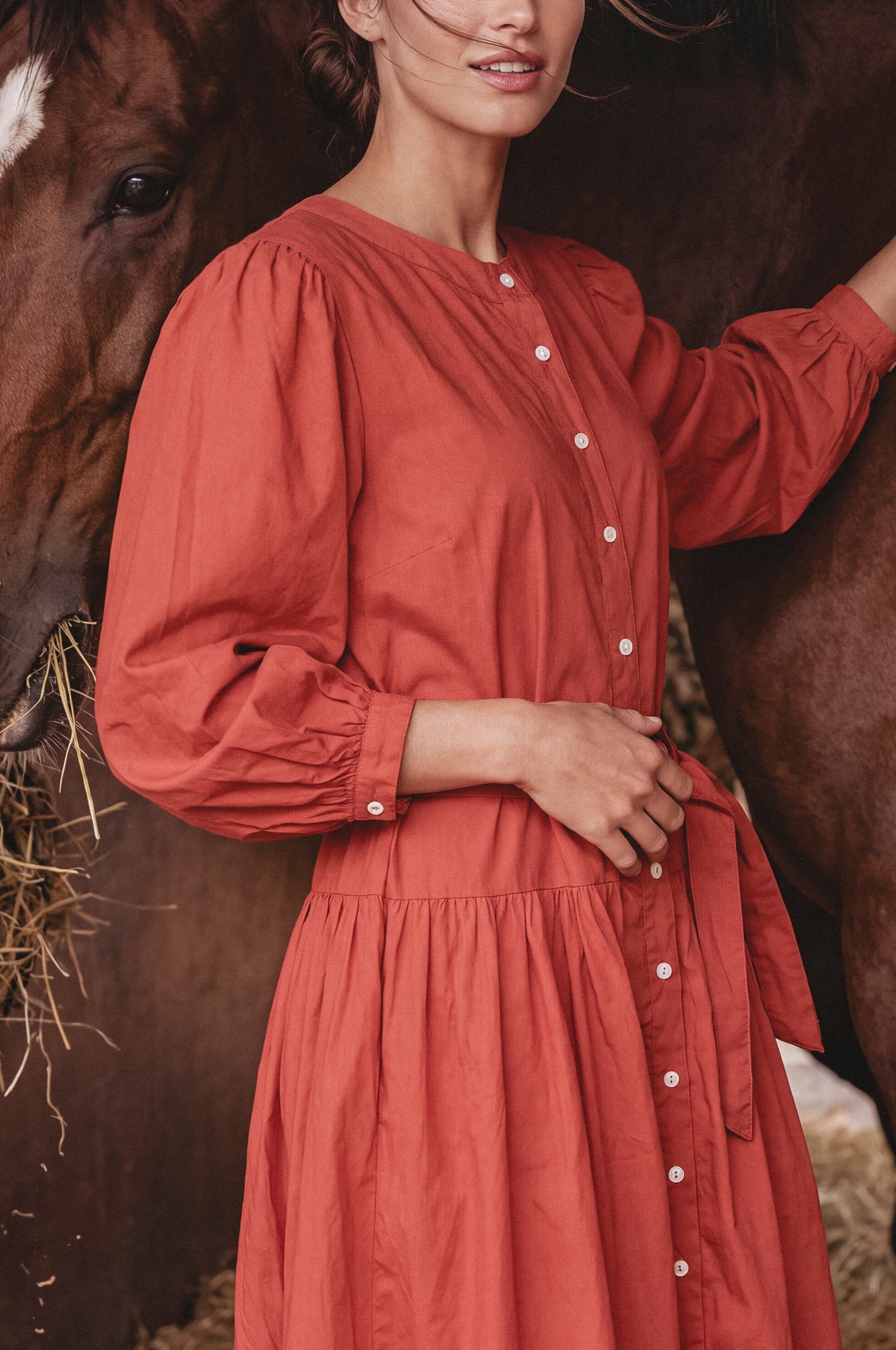 A woman wearing a rust-orange long-sleeve button-front dress stands beside a horse in a barn setting, with hay visible on the ground.