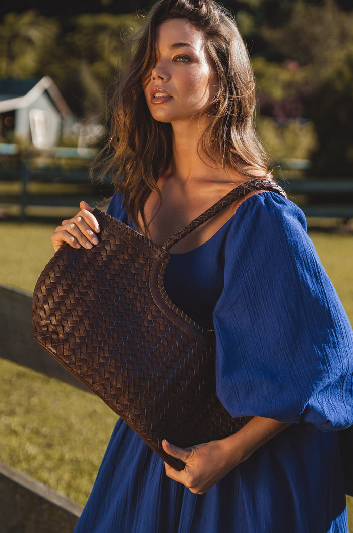 Woman in a blue dress holding a woven brown bag outdoors.