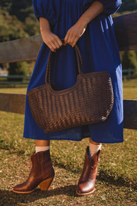 Person wearing a blue dress and brown boots holding a woven brown bag outdoors.