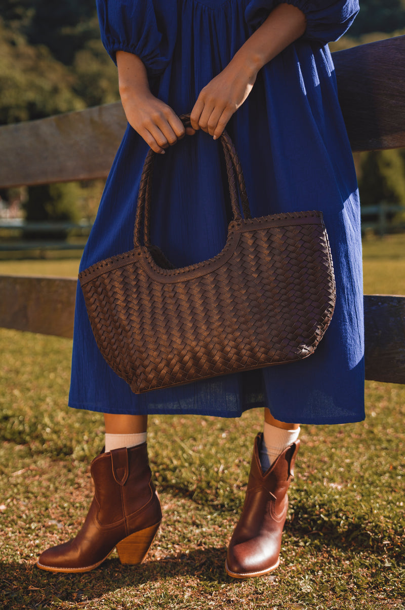 Person wearing a blue dress and brown boots holding a woven brown bag outdoors.