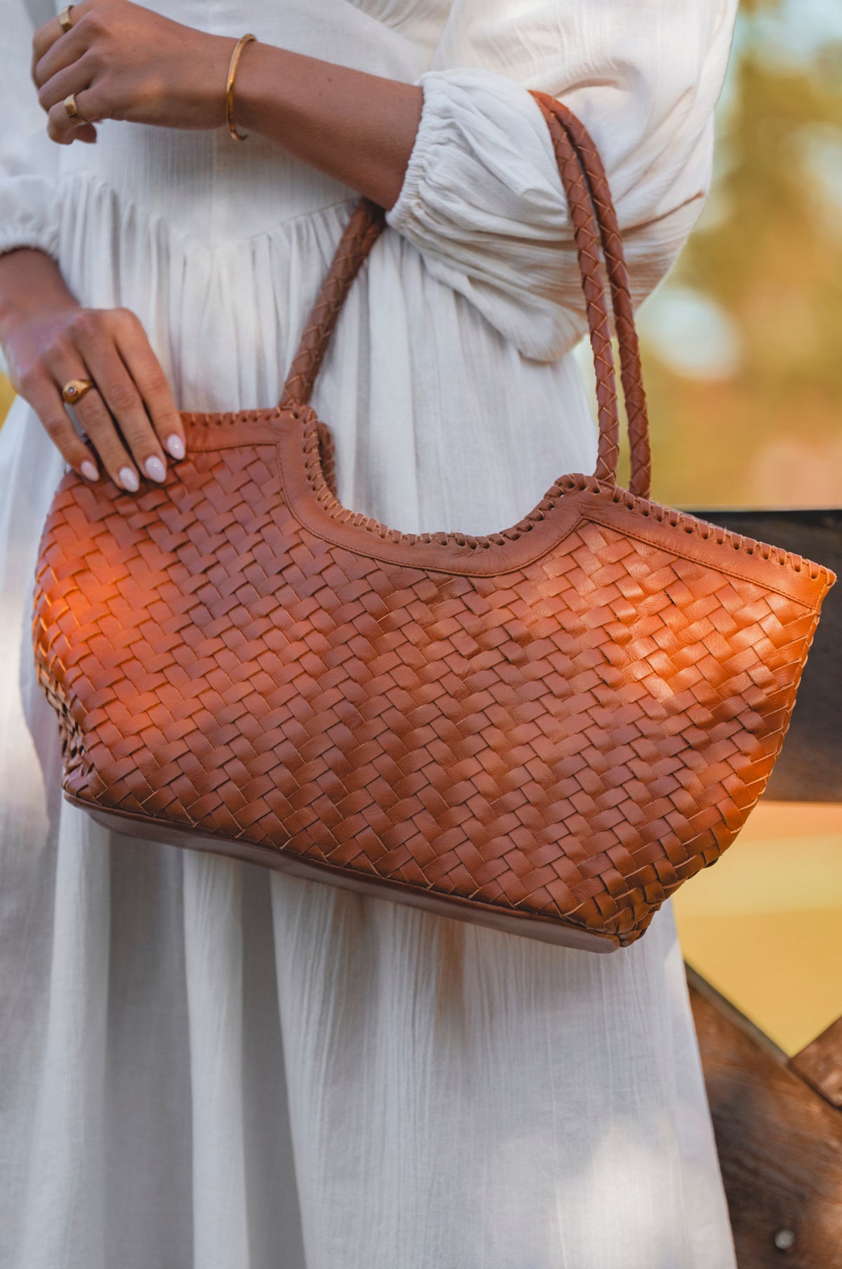 Brown woven handbag held by a person wearing a white dress.