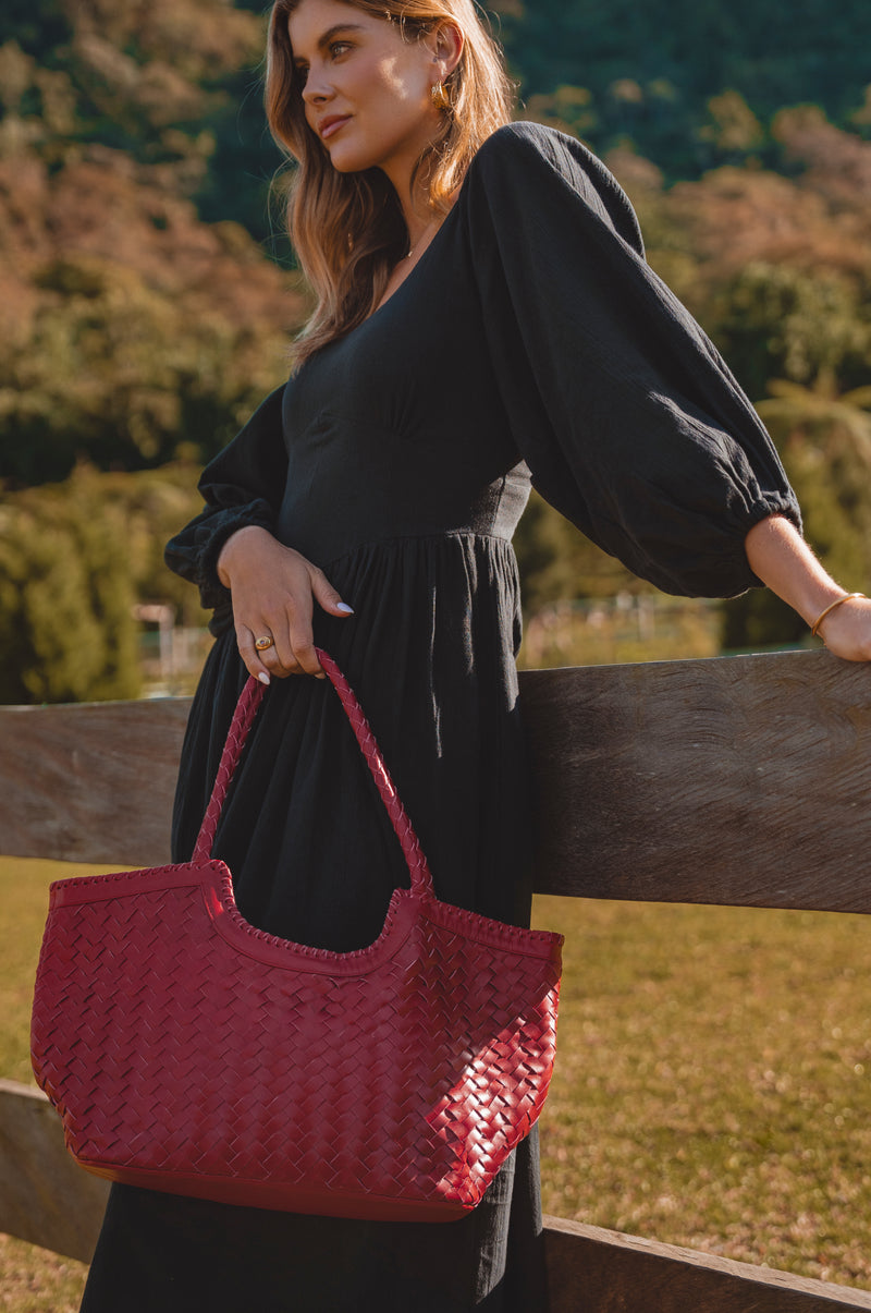 Woman holding a red textured handbag outdoors with greenery in the background