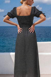 Woman in a black dress with puffed sleeves standing against a blue ocean and clear sky.