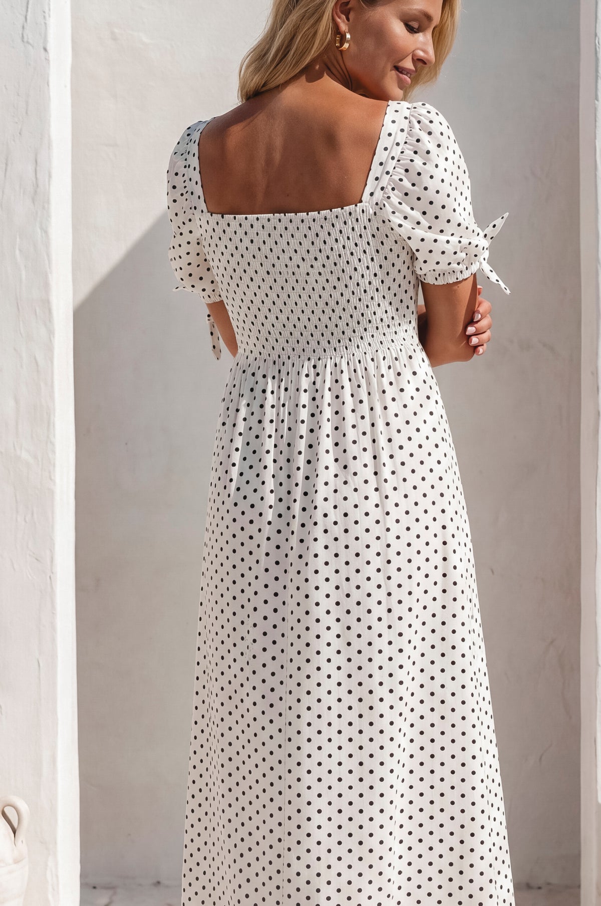 Woman wearing a white dress with black polka dots against a light wall.