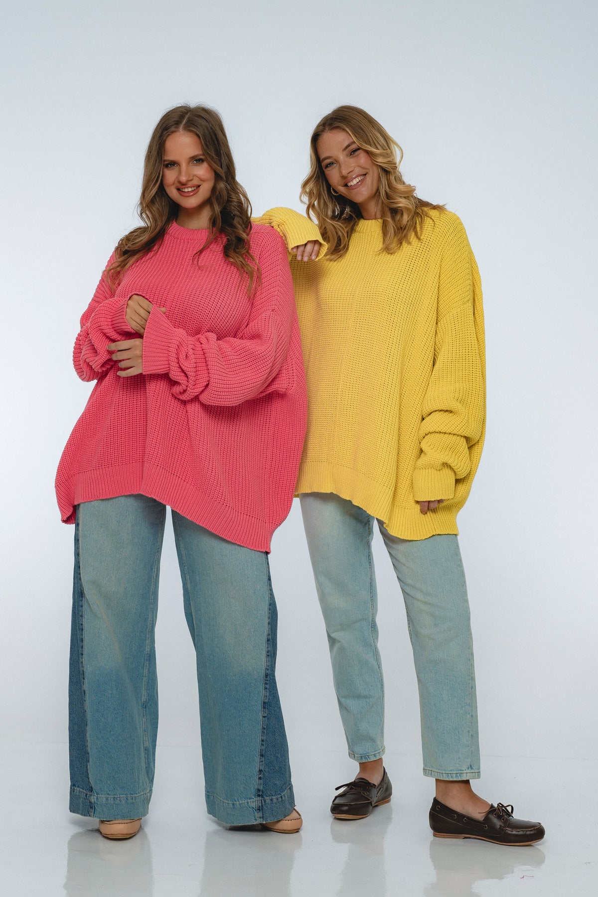 Two women wearing colorful sweaters and jeans on a white background
