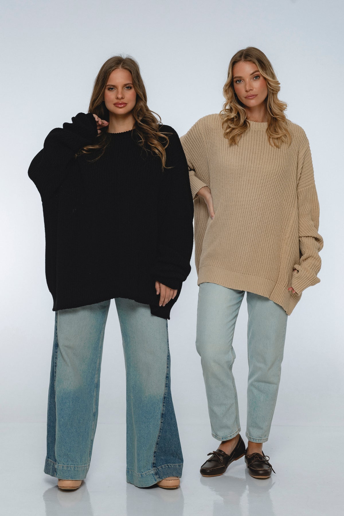 Two women wearing oversized sweaters and jeans on a white background