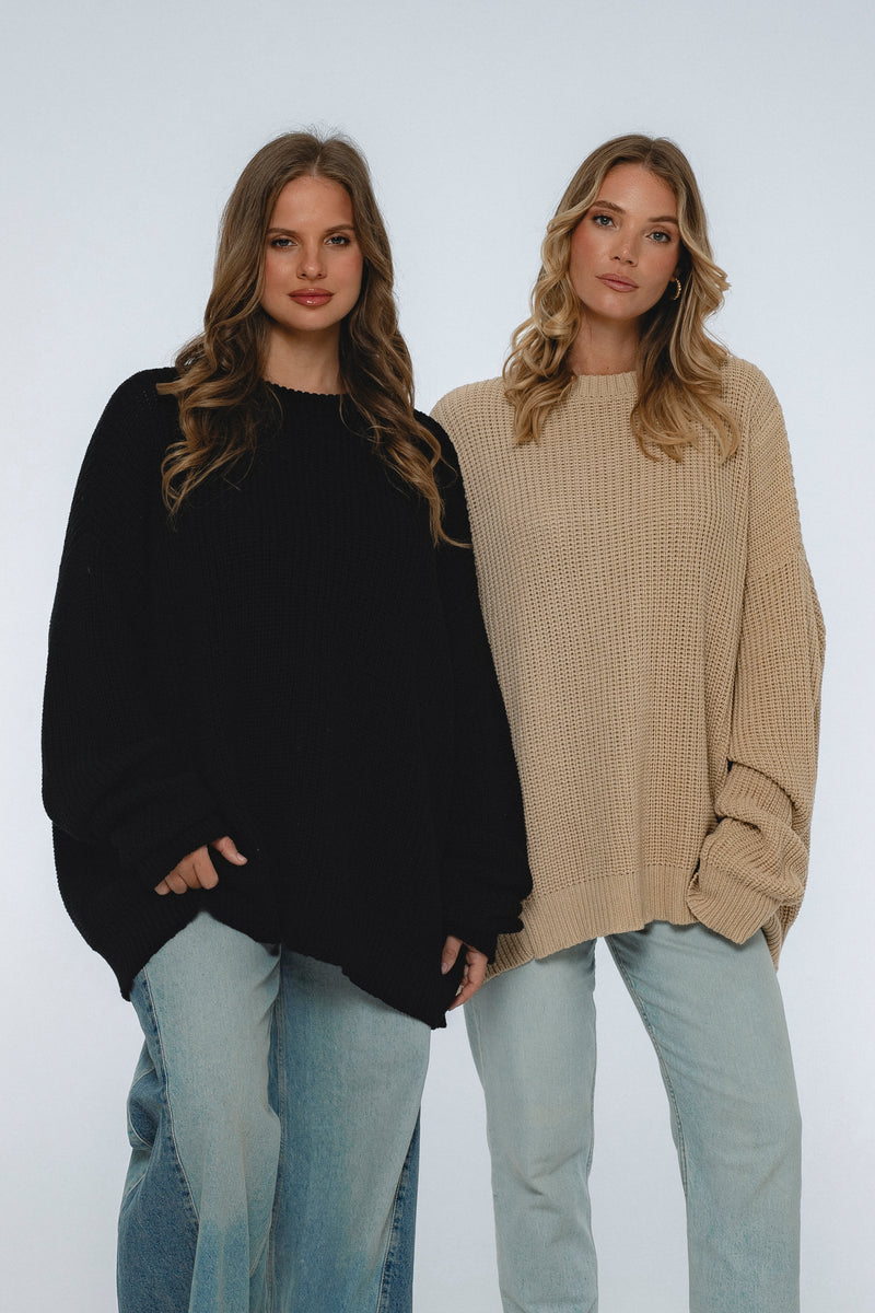 Two women wearing oversized sweaters and jeans against a white background