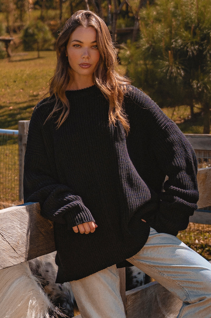 Woman wearing a black sweater and light jeans sitting outdoors.