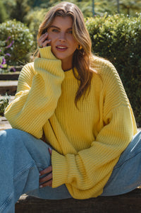 Woman wearing a yellow sweater sitting outdoors with greenery in the background
