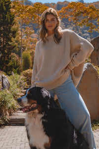 Woman in a beige sweater and jeans sitting next to a Bernese Mountain Dog in a park with trees and rocks.