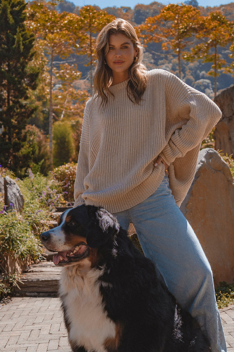 Woman in a beige sweater and jeans sitting next to a Bernese Mountain Dog in a park with trees and rocks.