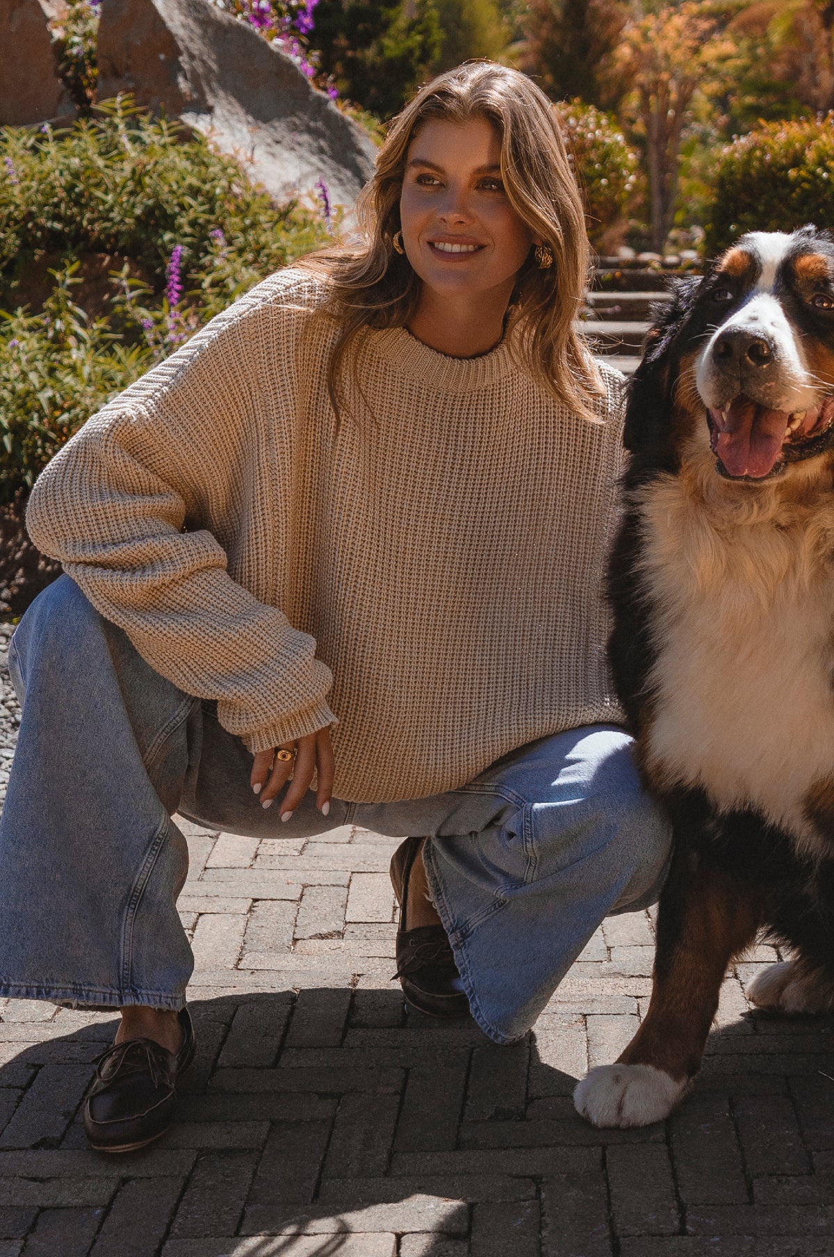 Woman in a beige sweater and jeans kneeling next to a Bernese Mountain Dog on a brick path.