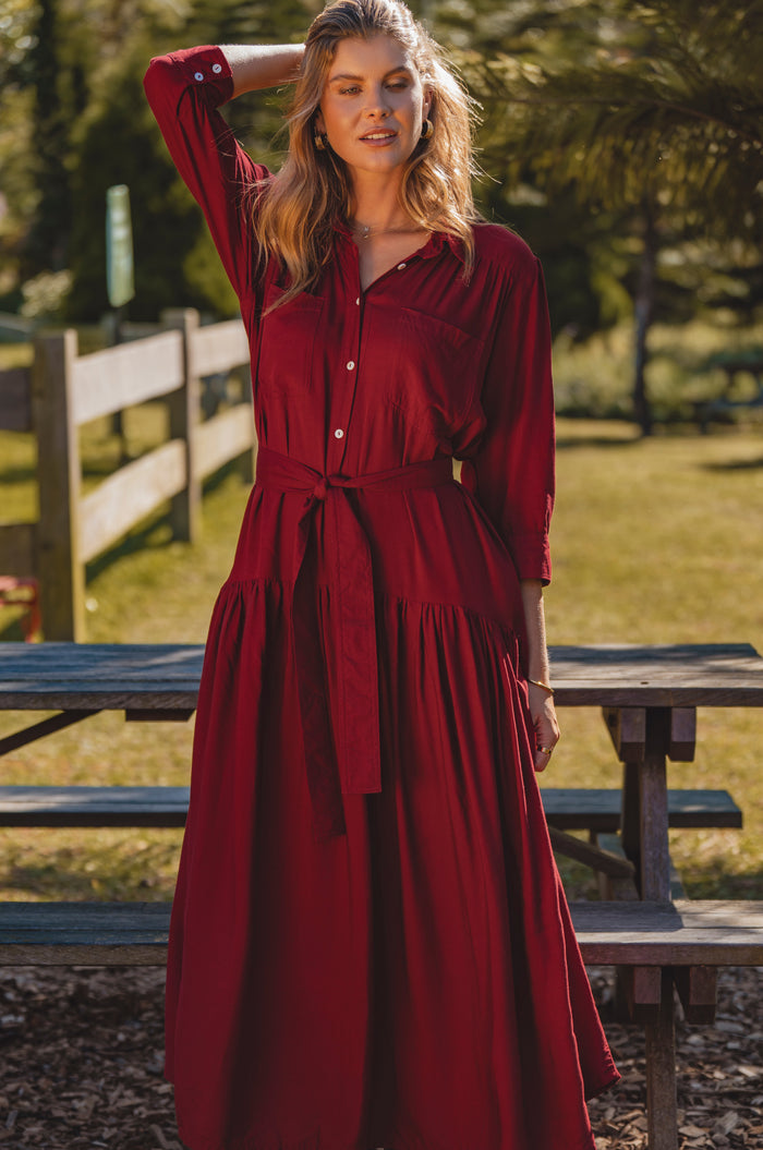 Woman wearing a red dress standing outdoors near a wooden bench.