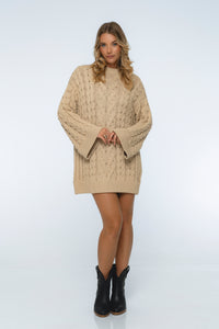 Woman wearing a beige cable knit sweater dress with black boots on a white background