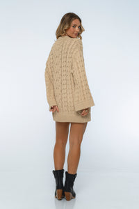 Woman wearing a beige cable knit cardigan over a beige dress on a white background