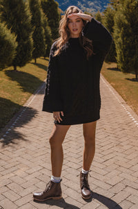 Woman in a black dress and brown boots standing on a paved path with trees in the background