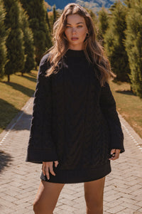 Woman wearing a black sweater dress in a scenic outdoor setting with trees and mountains.