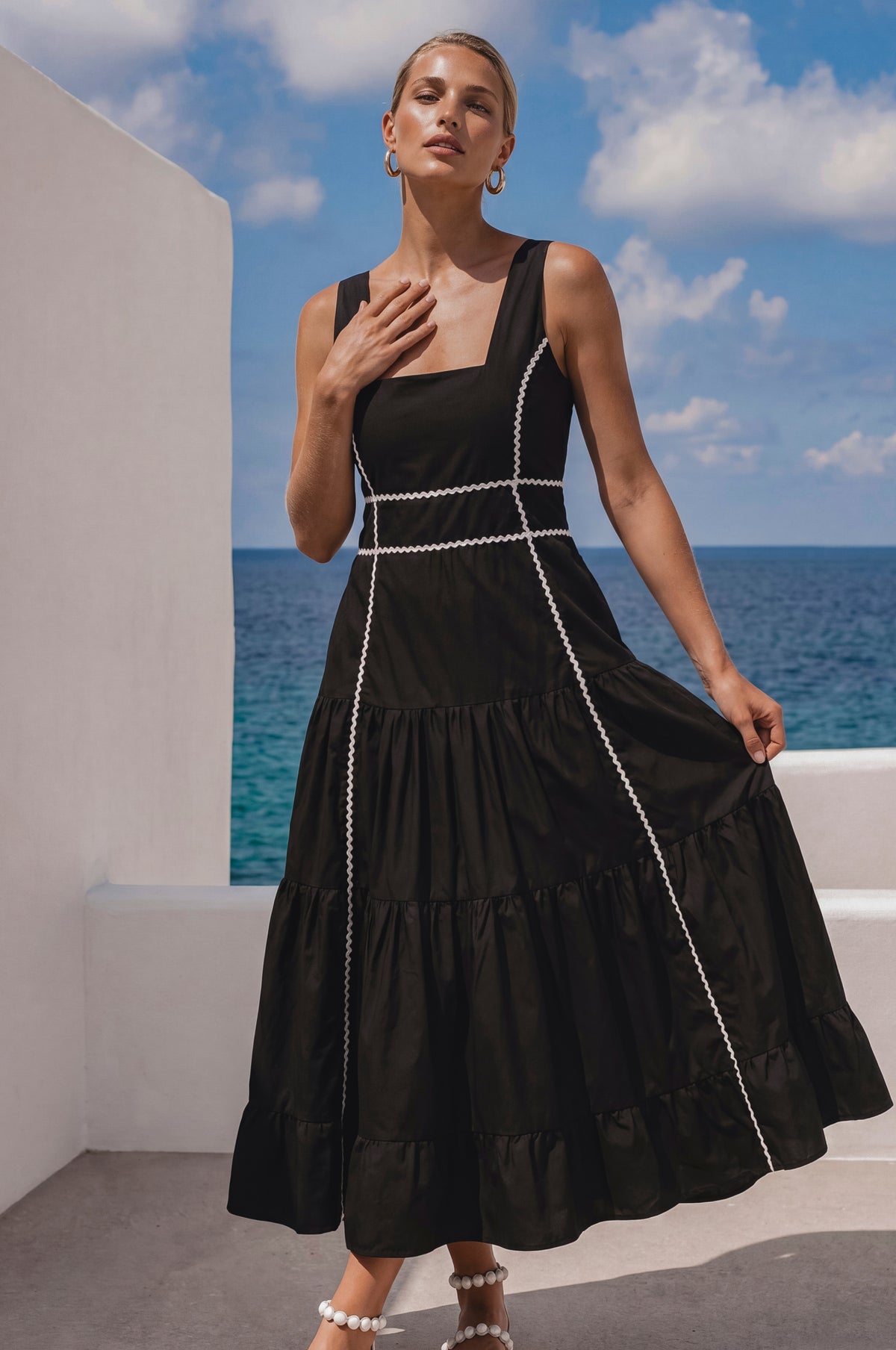 Woman in a black dress with white trim standing against a blue sky and ocean.