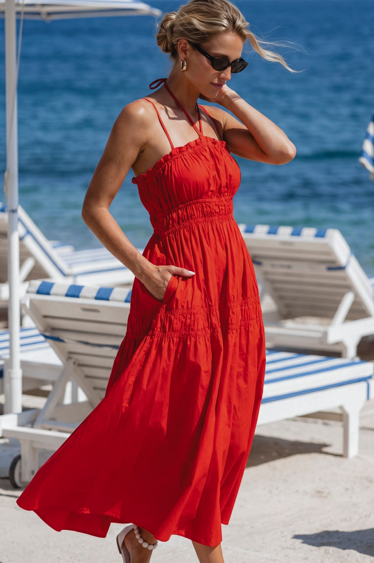 Woman in a red dress standing on a beach with blue ocean and white lounge chairs.
