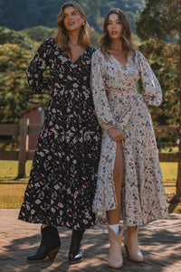 Two women wearing floral dresses standing outdoors with a wooden fence and greenery in the background.