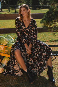 Woman in a floral dress sitting outdoors with a wooden cart of vegetables.