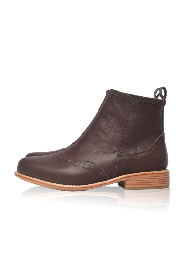 A dark brown leather ankle boot with a low stacked wood-look heel and a pull tab at the back, shown from a side profile on a white background with a soft reflection.