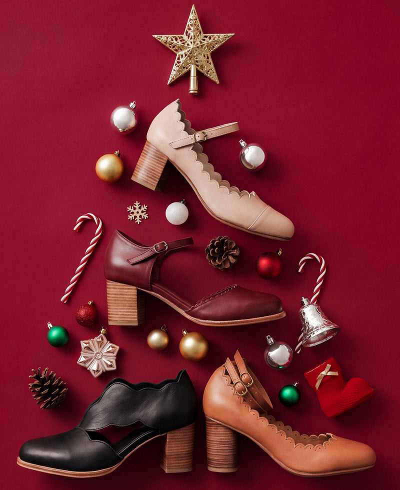 Four women's heeled shoes arranged in a Christmas tree shape on a deep red background, surrounded by holiday ornaments, candy canes, pinecones, and a gold star. The shoes include black, burgundy, tan, and beige styles with scalloped edges and block heels.