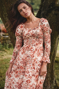 A woman wearing a long-sleeved bohemian-style floral dress leans against a tree outdoors. The dress features a warm cream base with rust red, terracotta, and orange floral patterns.