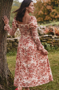 A woman wearing a long-sleeved midi dress with a red and pink floral pattern on a cream background, standing outdoors next to a tree.