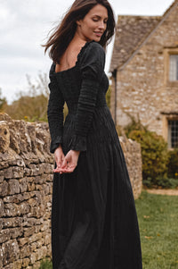 A woman wearing a long black smocked dress stands outdoors beside a stone wall and stone building, looking down and to the side.