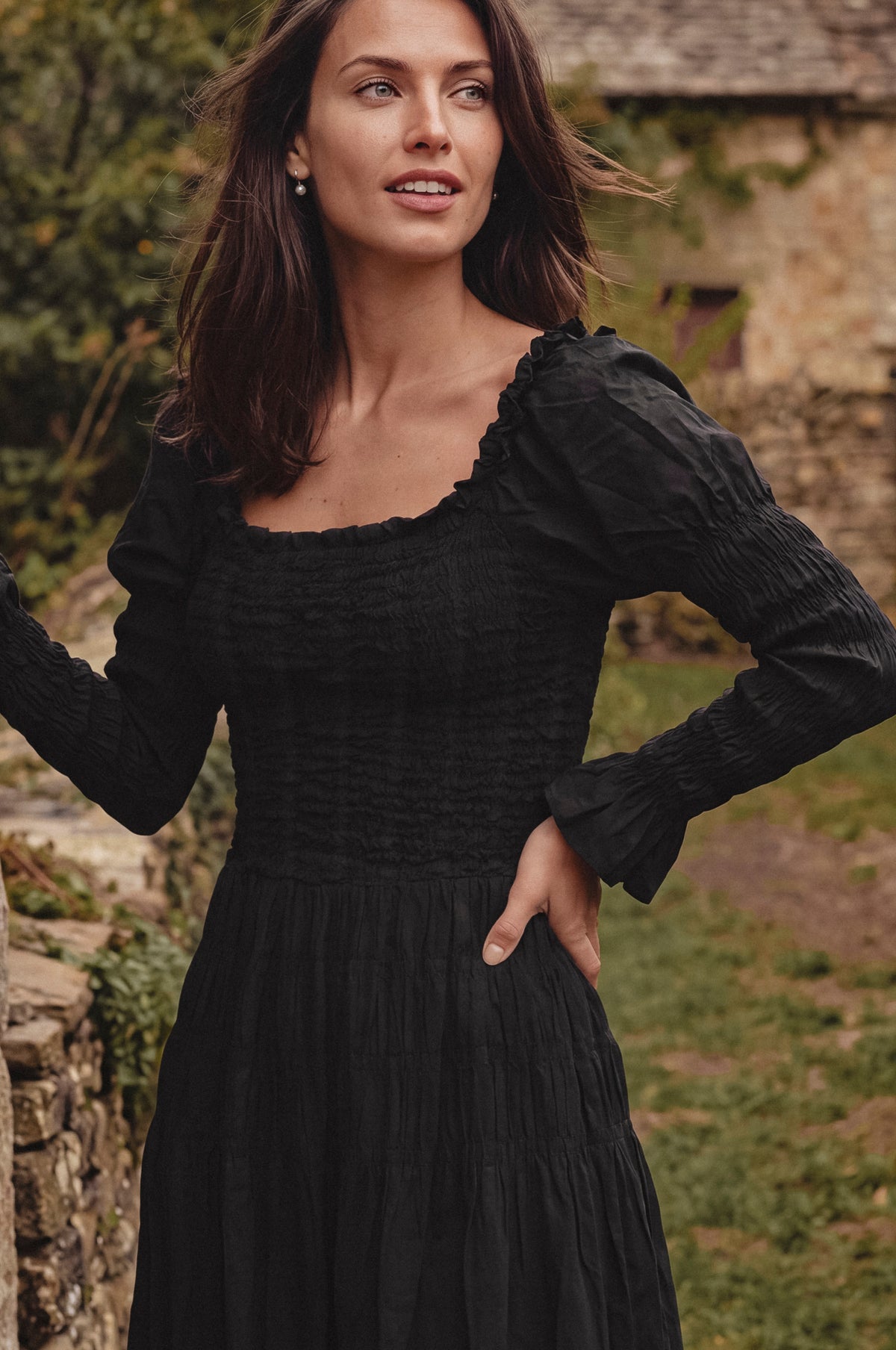 A woman wearing a black long-sleeve smocked dress, standing outdoors in front of a rustic stone wall and greenery.