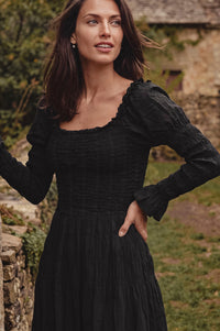 A woman wearing a black long-sleeve smocked dress, standing outdoors in front of a rustic stone wall and greenery.