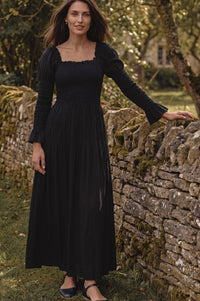 A woman wearing a long black midi dress with puff sleeves and a smocked bodice, standing outdoors next to a mossy stone wall.