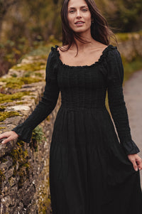 A woman wearing a long-sleeved black midi dress with a smocked bodice and ruffled neckline, standing outdoors next to a mossy stone wall.