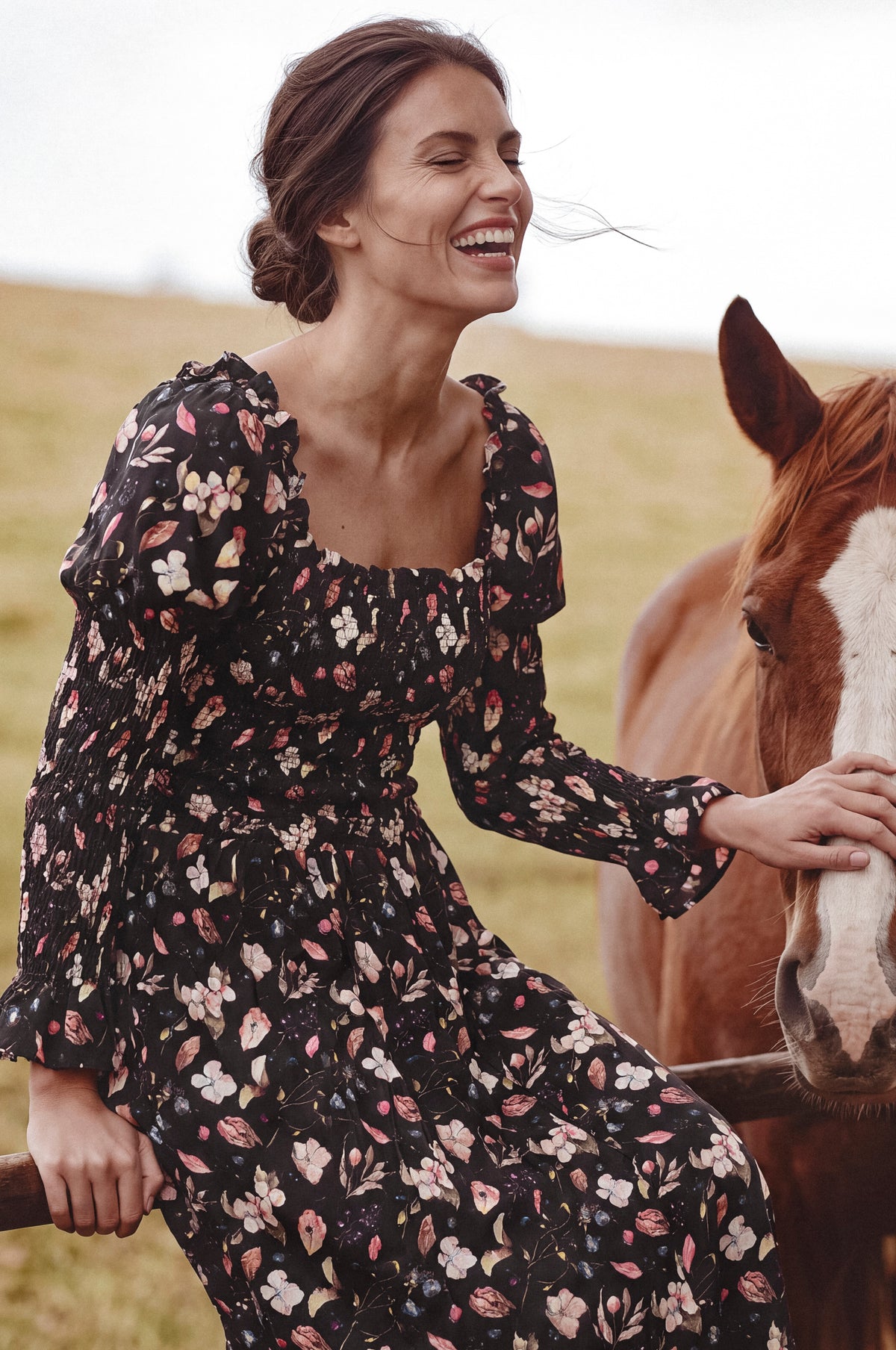 A woman wearing a black floral long-sleeve dress sits beside a brown horse in an outdoor field. The dress has a black base with a multicolor floral pattern in pink, white, and light blue.