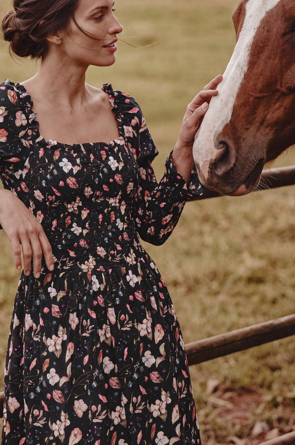A woman in a black floral long-sleeve dress stands outdoors next to a brown and white horse, gently touching its face in a grassy field.
