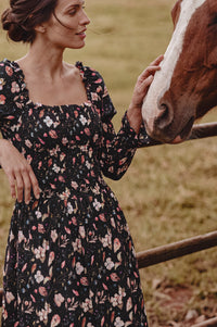 A woman in a black floral long-sleeve dress stands outdoors next to a brown and white horse, gently touching its face in a grassy field.