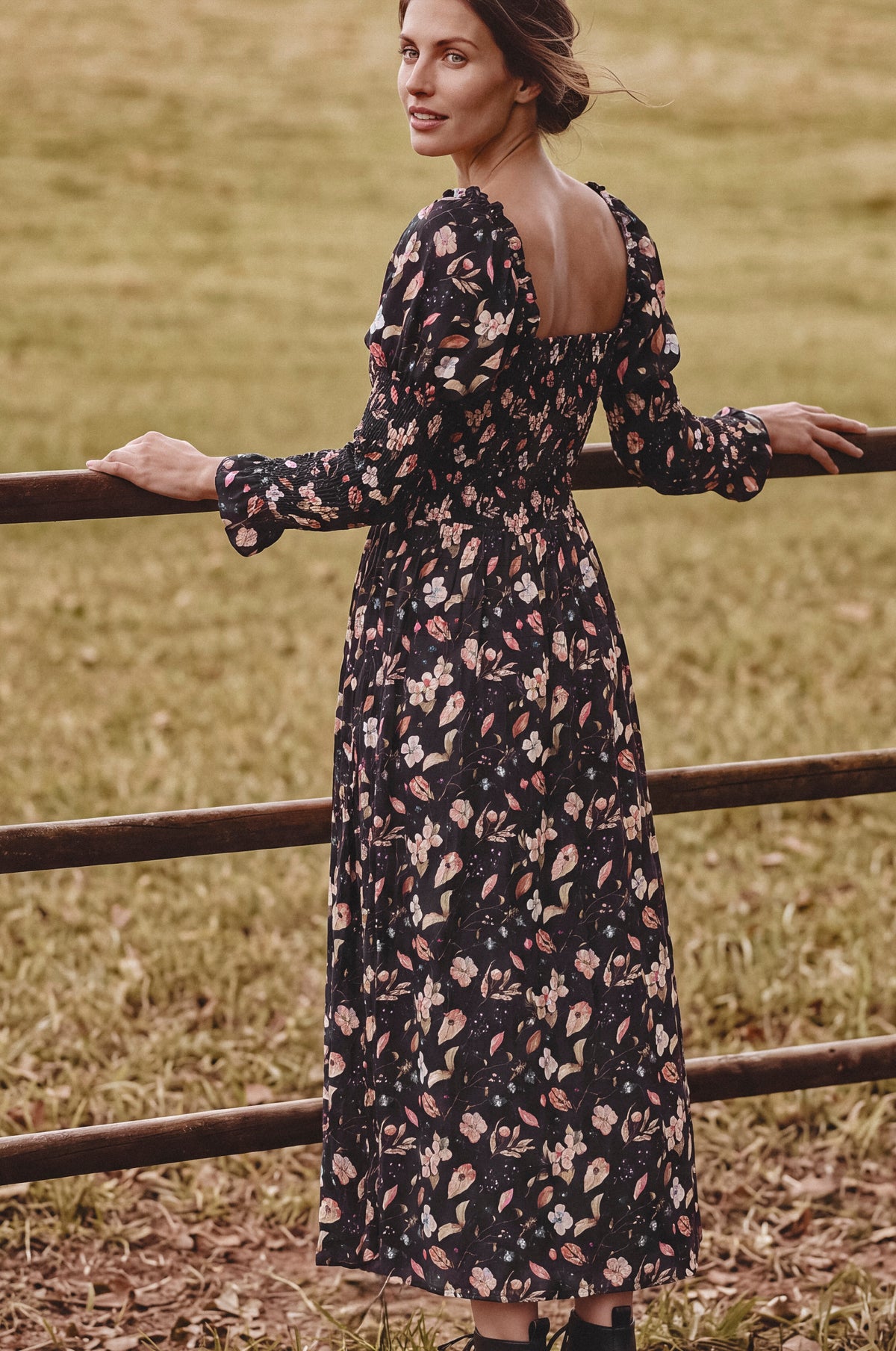 A woman wearing a long-sleeve black midi dress with a pink and beige floral print, standing outdoors in a grassy field next to a wooden fence.