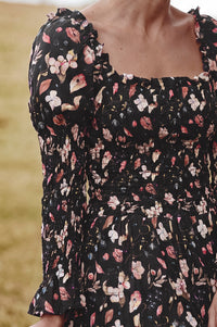 Close-up of a woman wearing a black smocked dress with a pink, white, and beige floral print, featuring puff sleeves and a square neckline.