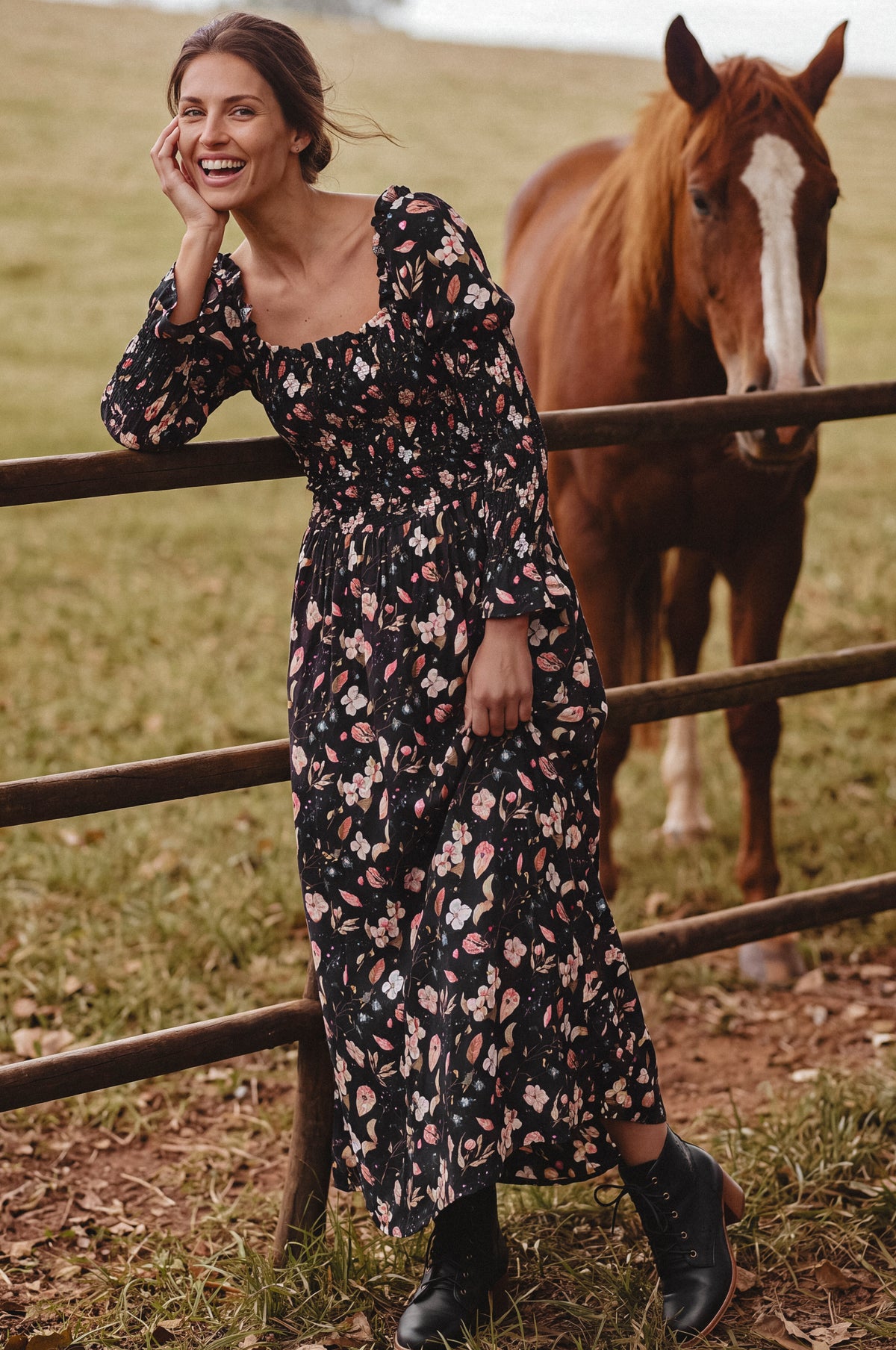 A woman in a black floral midi dress leans against a wooden fence in a grassy field, with a brown horse standing behind the fence.