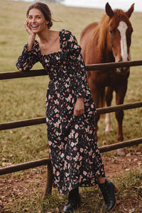 A woman in a black floral midi dress leans against a wooden fence in a grassy field, with a brown horse standing behind the fence.