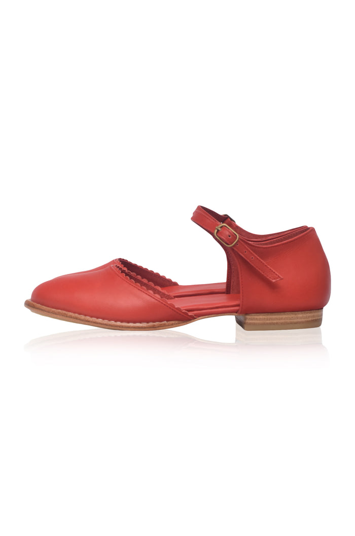 A red leather Mary Jane flat shoe with a strap across the instep, a small gold-tone buckle, and a low stacked heel, shown from a side profile on a white background.