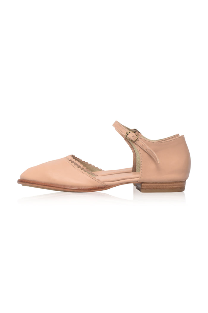 A single light beige leather flat shoe with a T-strap and low block heel, shown from a side profile on a white background.