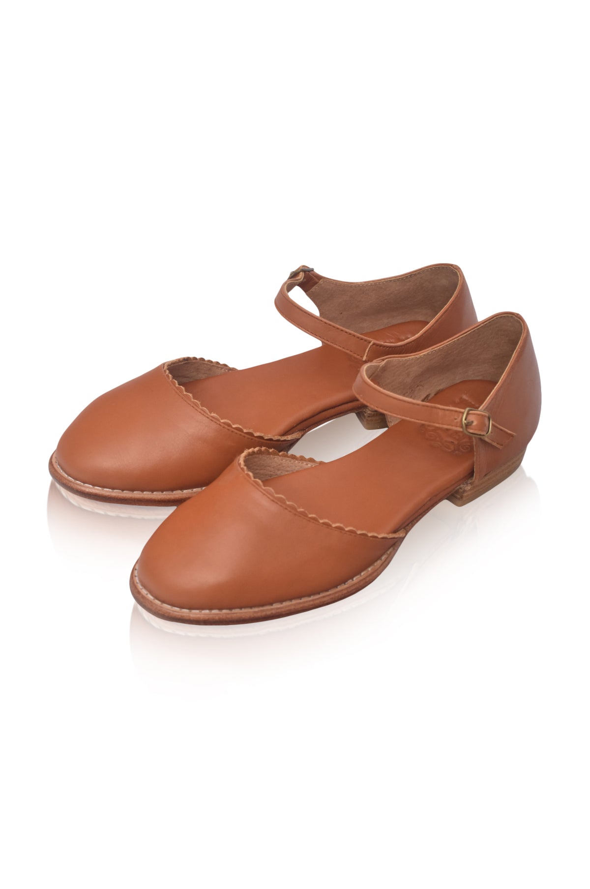A pair of women's low-heeled flat shoes in a warm tan-brown leather, featuring a rounded toe, decorative scalloped edge stitching, and a single ankle strap with a small gold-tone buckle, shown on a white background with a soft reflection.